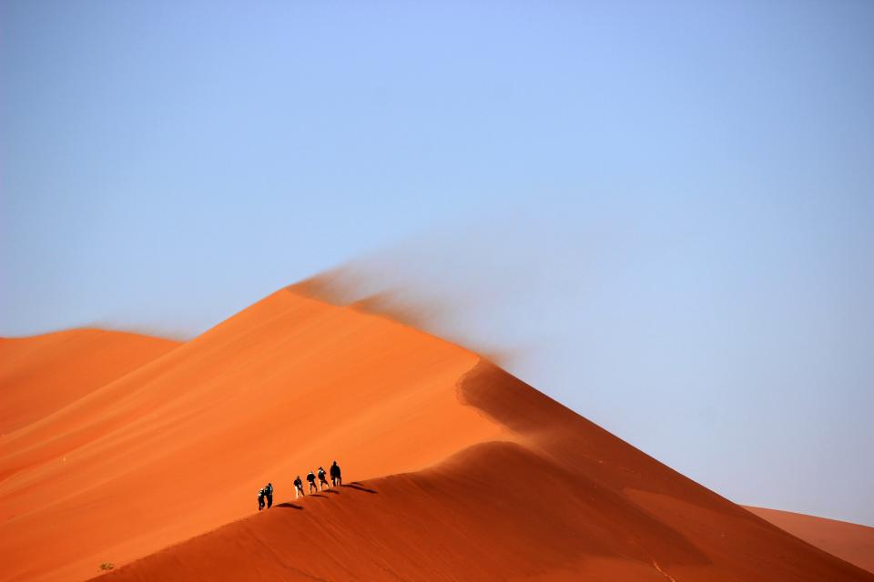 windy trek sunny sky sanddunes people orange hot hills hiking hike desert blue 
